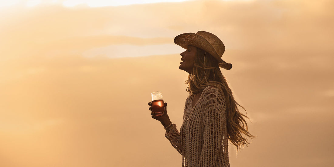A woman in a hat holding a drink against a sunset background.